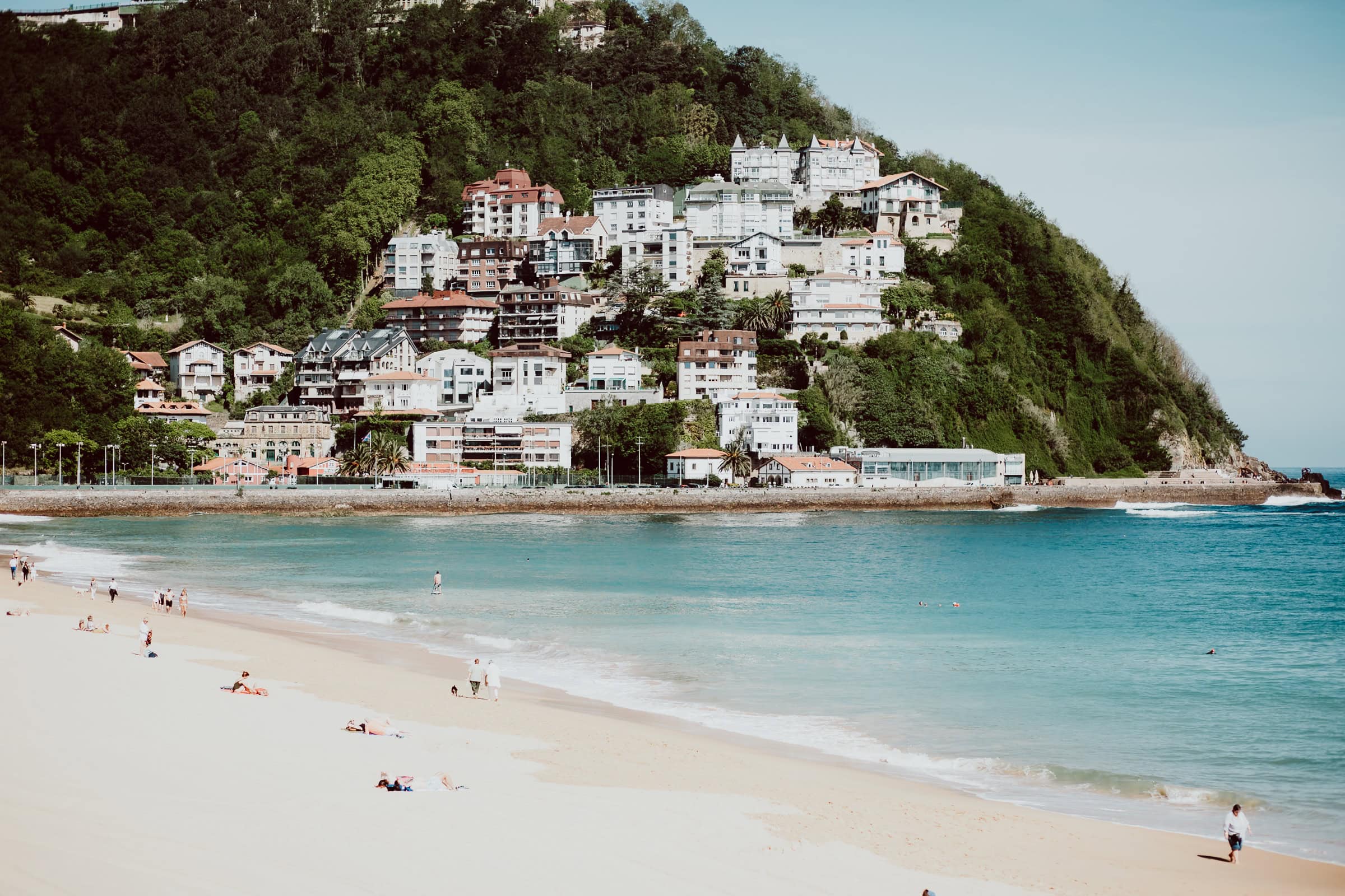 Strand mit Gebäude und grüner Hangkulisse.